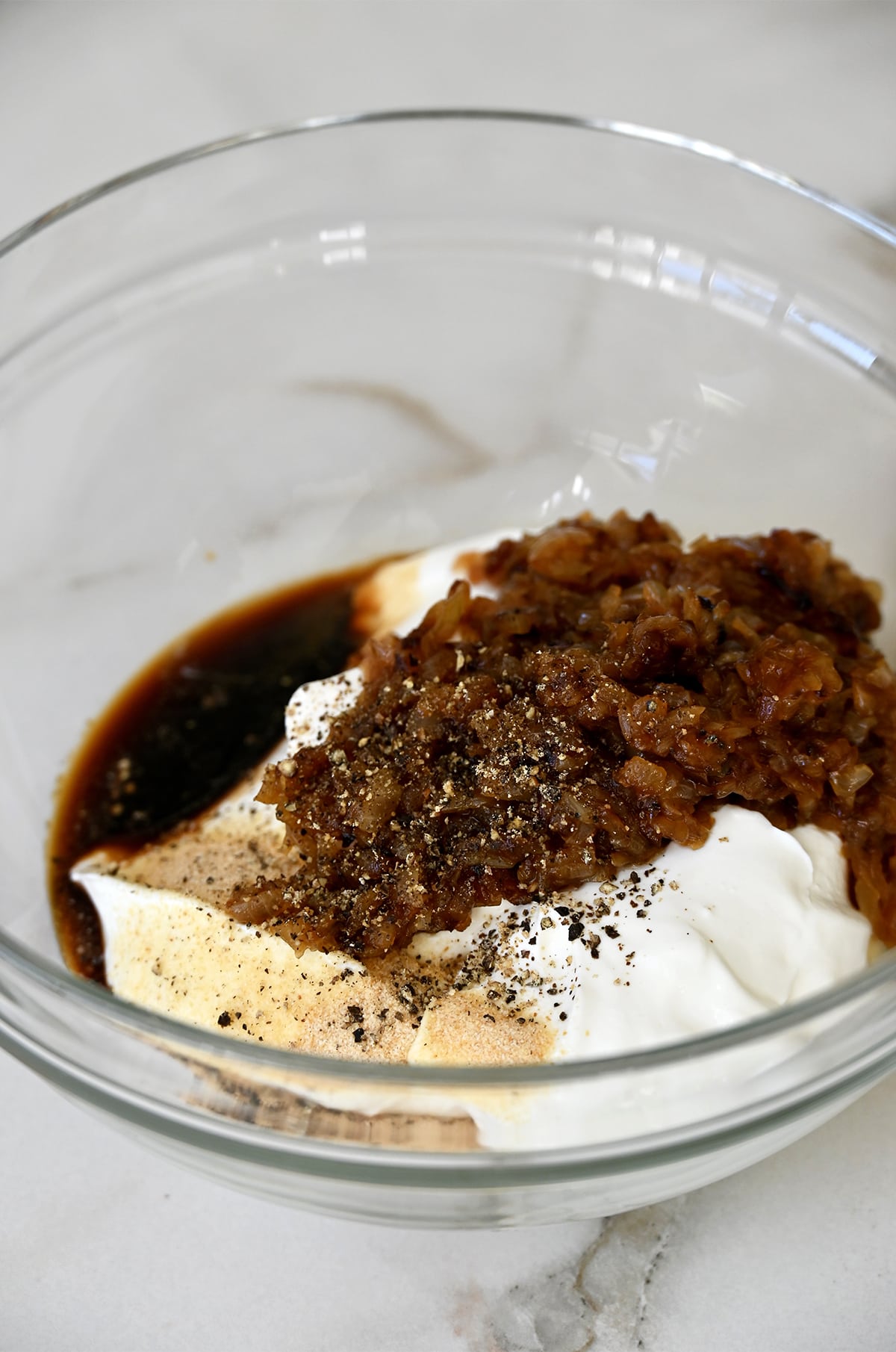 A glass bowl containing sour cream, soy sauce, caramelized onions, garlic powder and black pepper.