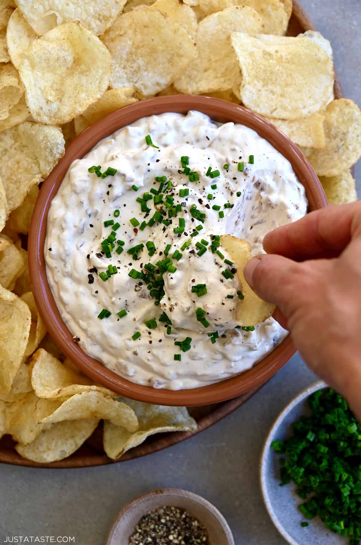A hand holding a potato chip dips it into a bowl containing sour cream and onion dip that's on a plate surrounded by potato chips.
