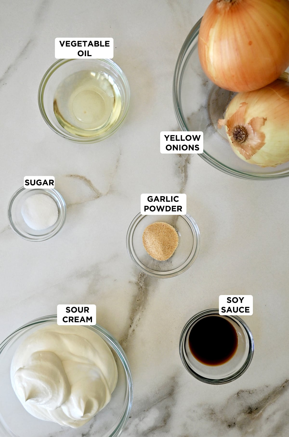 Clear glass bowls containing the ingredients for French onion dip, including two yellow onions, vegetable oil, garlic powder, soy sauce, sour cream and sugar.