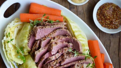 White plate containing The Best Slow Cooker Corned Beef and Cabbage with carrots next to a loaf of bread and two small bowls containing mustards