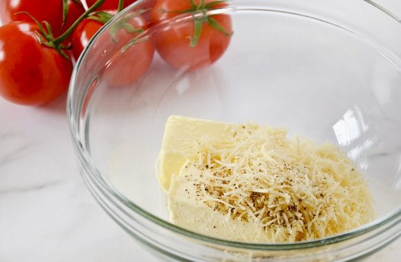 A glass bowl containing butter, shredded Parmesan cheese, minced garlic and pepper with vine-ripe tomatoes in background