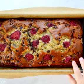 A baked loaf of Raspberry Chocolate Chip Banana Bread with a child's hand touching it