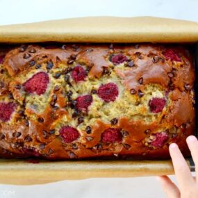 A baked loaf of Raspberry Chocolate Chip Banana Bread with a child's hand touching it