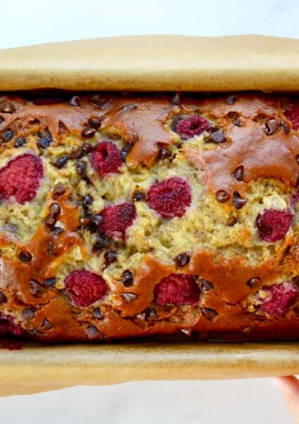 A baked loaf of Raspberry Chocolate Chip Banana Bread with a child's hand touching it