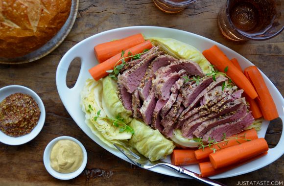 White serving plate with corned beef, cabbage and carrots next to a loaf of bread and two small dishes with mustard