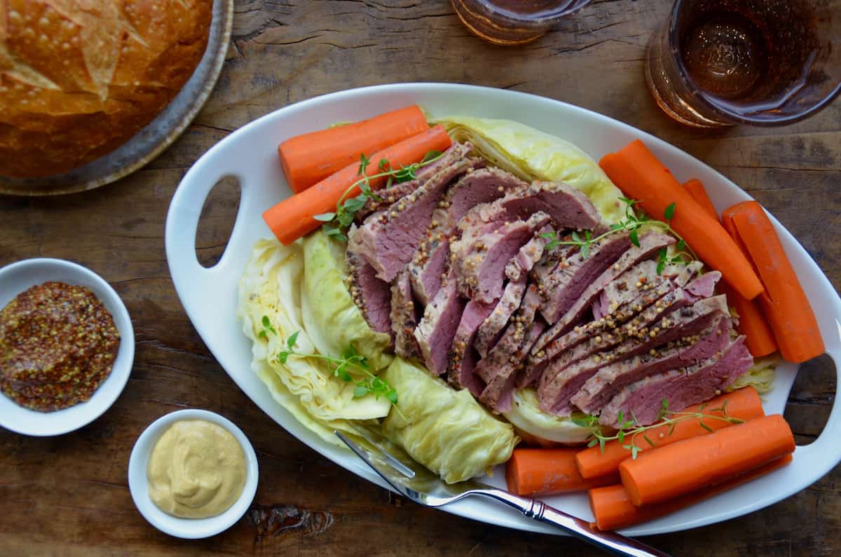 Sliced corned beef is surrounded with cabbage and carrots on an oval platter. Nearby are dishes of mustard and a loaf of soda bread.