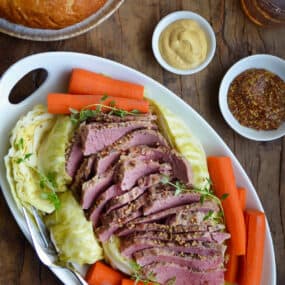 Sliced corned beef is surrounded with cabbage and carrots on an oval platter. Nearby are dishes of mustard and a loaf of soda bread.