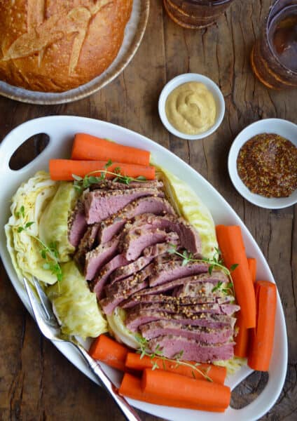 Sliced corned beef is surrounded with cabbage and carrots on an oval platter. Nearby are dishes of mustard and a loaf of soda bread.