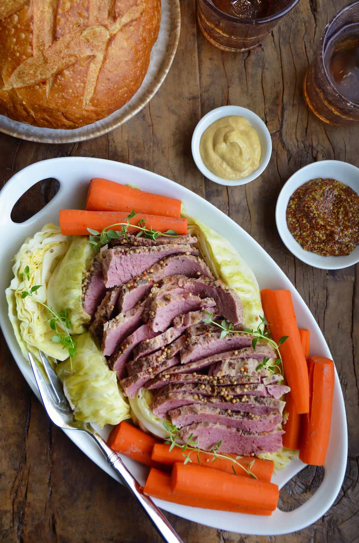 Sliced corned beef is surrounded with cabbage and carrots on an oval platter. Nearby are dishes of mustard and a loaf of soda bread.