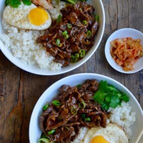 Top down view of two bowls containing easy beef bulgogi, white rice, a fried egg, cucumber ribbons, fresh cilantro and kimchi