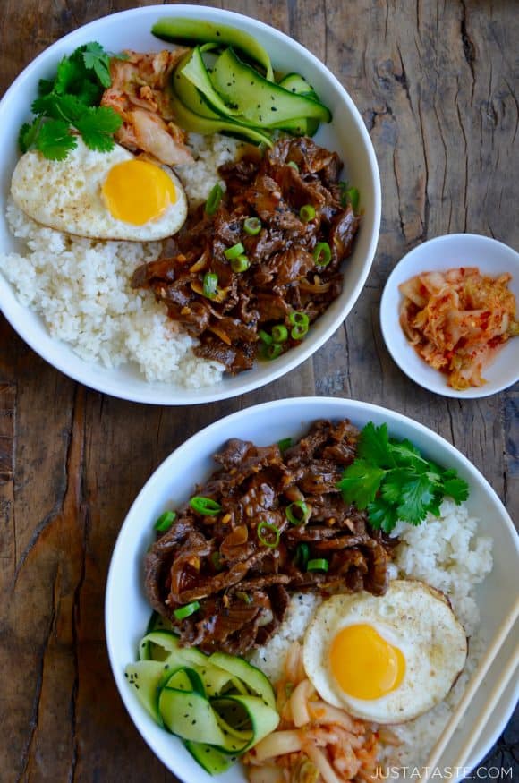 Top down view of two bowls containing easy beef bulgogi, white rice, a fried egg, cucumber ribbons, fresh cilantro and kimchi 