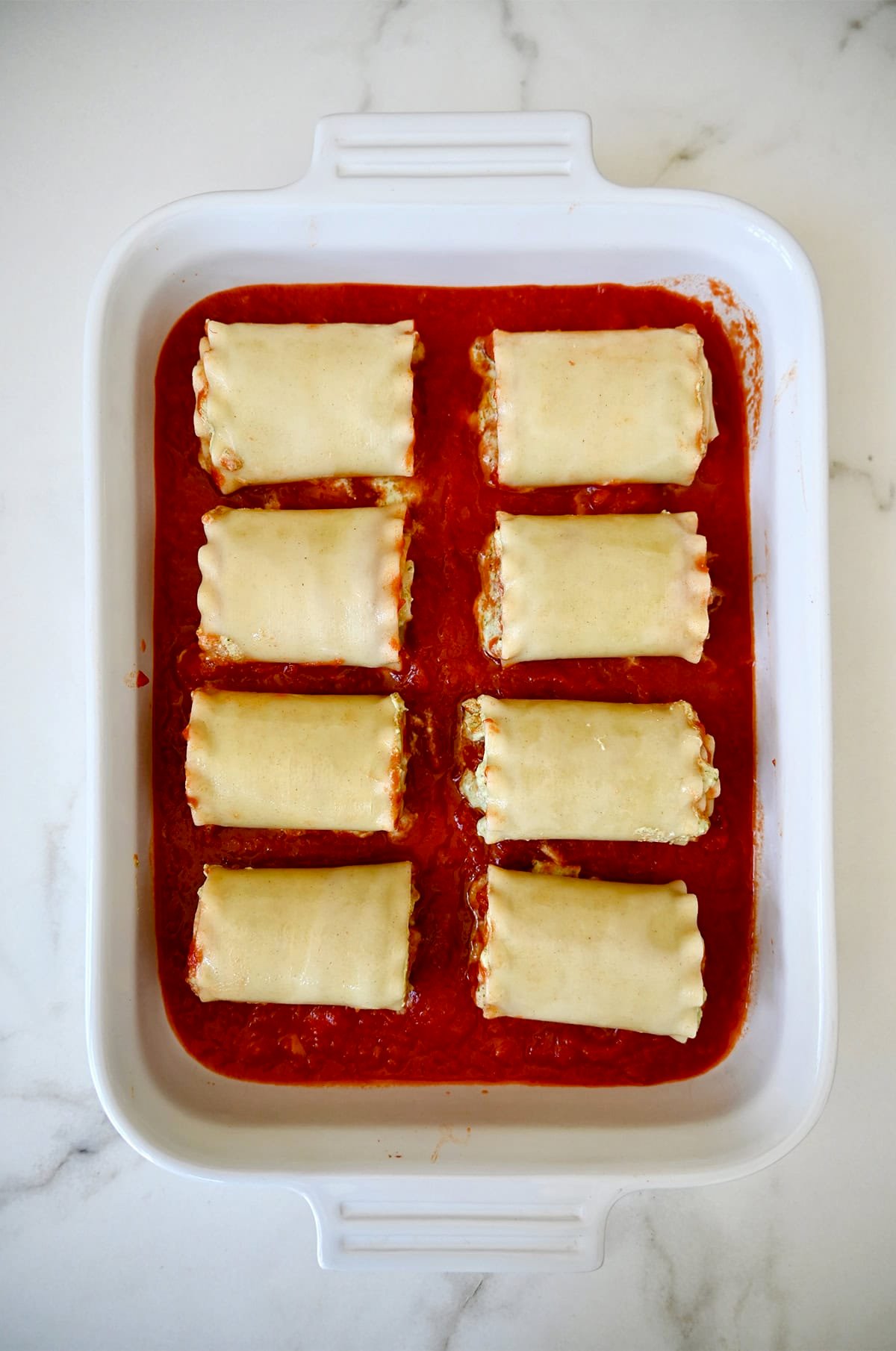 Lasagna roll-ups atop marinara sauce in a white baking dish. 