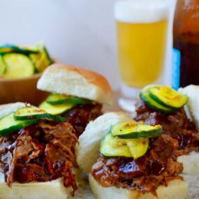 White buns piled high with Instant Pot Barbecue Pulled Pork and pickles in front of a bottle and glass of beer