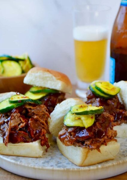White buns piled high with Instant Pot Barbecue Pulled Pork and pickles in front of a bottle and glass of beer