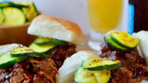 White buns piled high with Instant Pot Barbecue Pulled Pork and pickles in front of a bottle and glass of beer