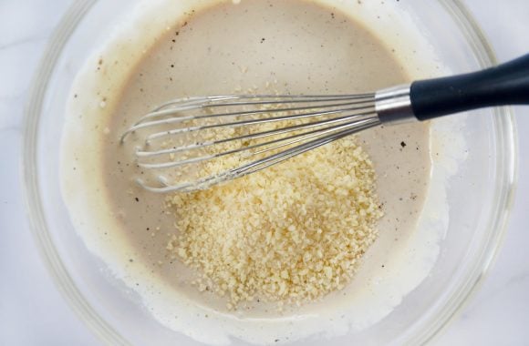 Glass bowl containing caesar salad dressing, parmesan cheese and a whisk