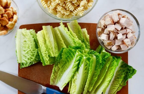 Chopped romaine on cutting board with knife next to three bowls containing chicken, pasta and croutons