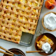 A top-down view of a peach slab pie with the corner piece on a plate with a dollop of whipped cream and a spoon.