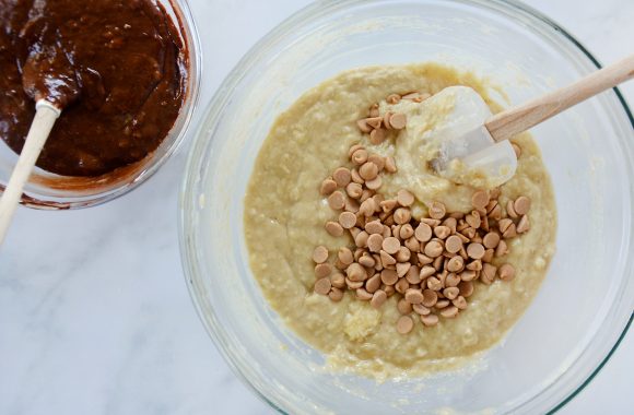 A small glass bowl containing chocolate banana bread batter with a spatula and a large glass bowl containing banana bread batter with peanut butter chips being stirred with a white spatula