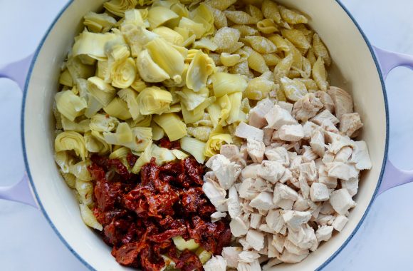 Top down view of large pot containing cooked pasta, chicken, sundried tomatoes and diced artichoke hearts