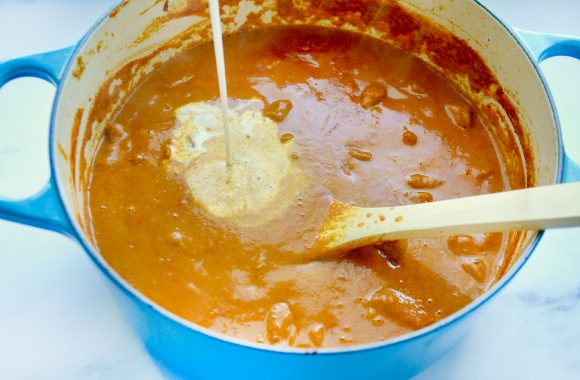 Heavy cream being added to a blue saucepot containing curry sauce and a wooden spoon 