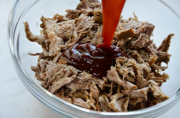 Clear bowl containing pulled pork with barbecue sauce being added 