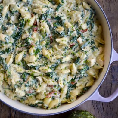 Top down view of a saucepot containing Spinach Artichoke Dip Pasta with Chicken next to an artichoke cut in half