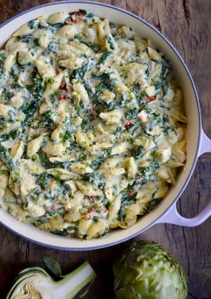 Top down view of a saucepot containing Spinach Artichoke Dip Pasta with Chicken next to an artichoke cut in half