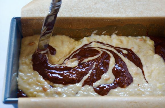 Knife swirling together banana bread batter and chocolate mixture in bread pan