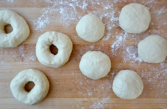 A wood cutting board containing unbaked, shaped bagels and balls of bagel dough