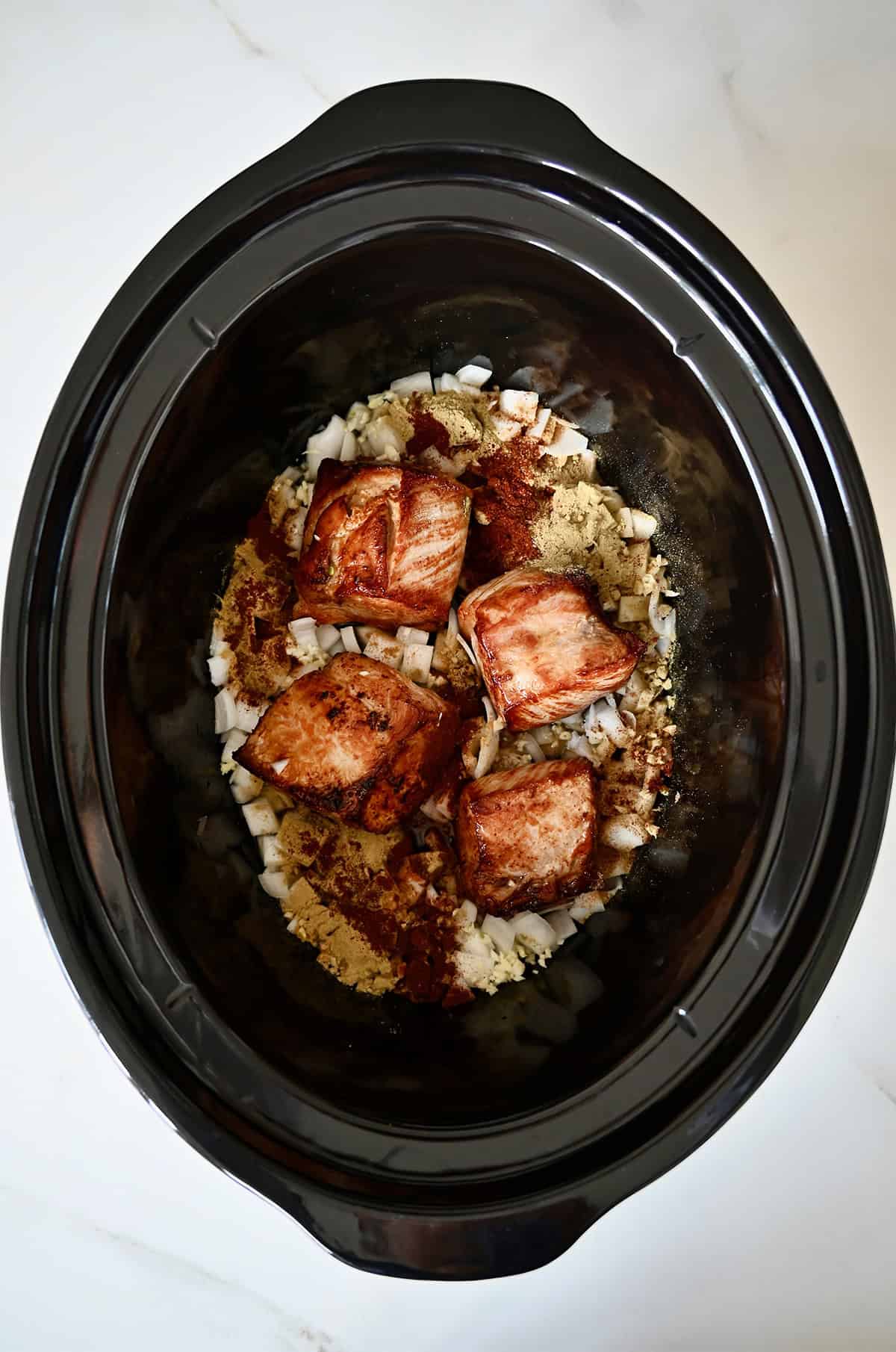 Adding pork shoulder to a slow cooker with spices, chicken stock and chopped onions to make carnitas.