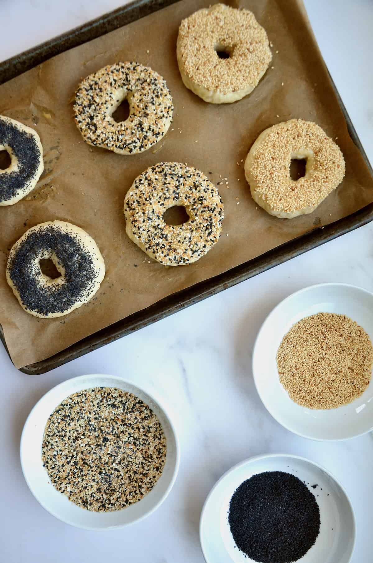Unbaked homemade bagels on a baking sheet with three different toppings: poppyseed, sesame and everything bagel.