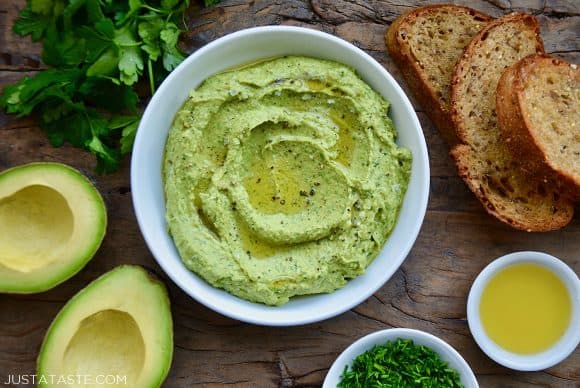 A white bowl containing avocado hummus surrounded by toast, herbs and olive oil