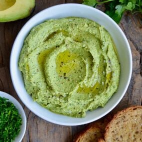 A white bowl containing avocado hummus with toast and herbs around it