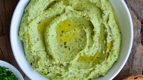 A white bowl containing avocado hummus with toast and herbs around it
