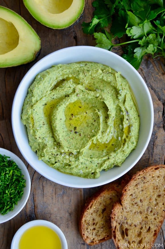 A white bowl containing avocado hummus with toast and herbs around it