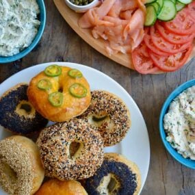 A white platter with homemade bagels surrounded by a lox platter and three bowls of cream cheese