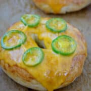 A close-up view of a cheddar-jalapeno bagel topped with a slice of cheddar cheese and slices of jalapeno pepper.