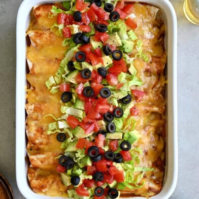 Rotisserie chicken enchiladas topped with shredded lettuce, black olives, diced tomatoes and avocado in a white baking dish.