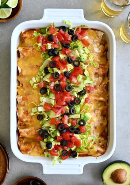 Rotisserie chicken enchiladas topped with shredded lettuce, black olives, diced tomatoes and avocado in a white baking dish.