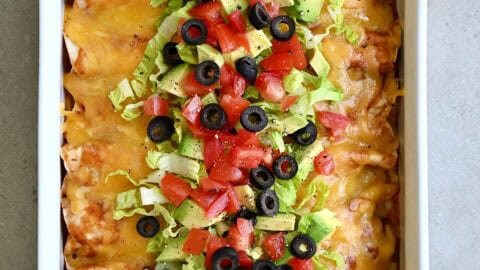 Rotisserie chicken enchiladas topped with shredded lettuce, black olives, diced tomatoes and avocado in a white baking dish.