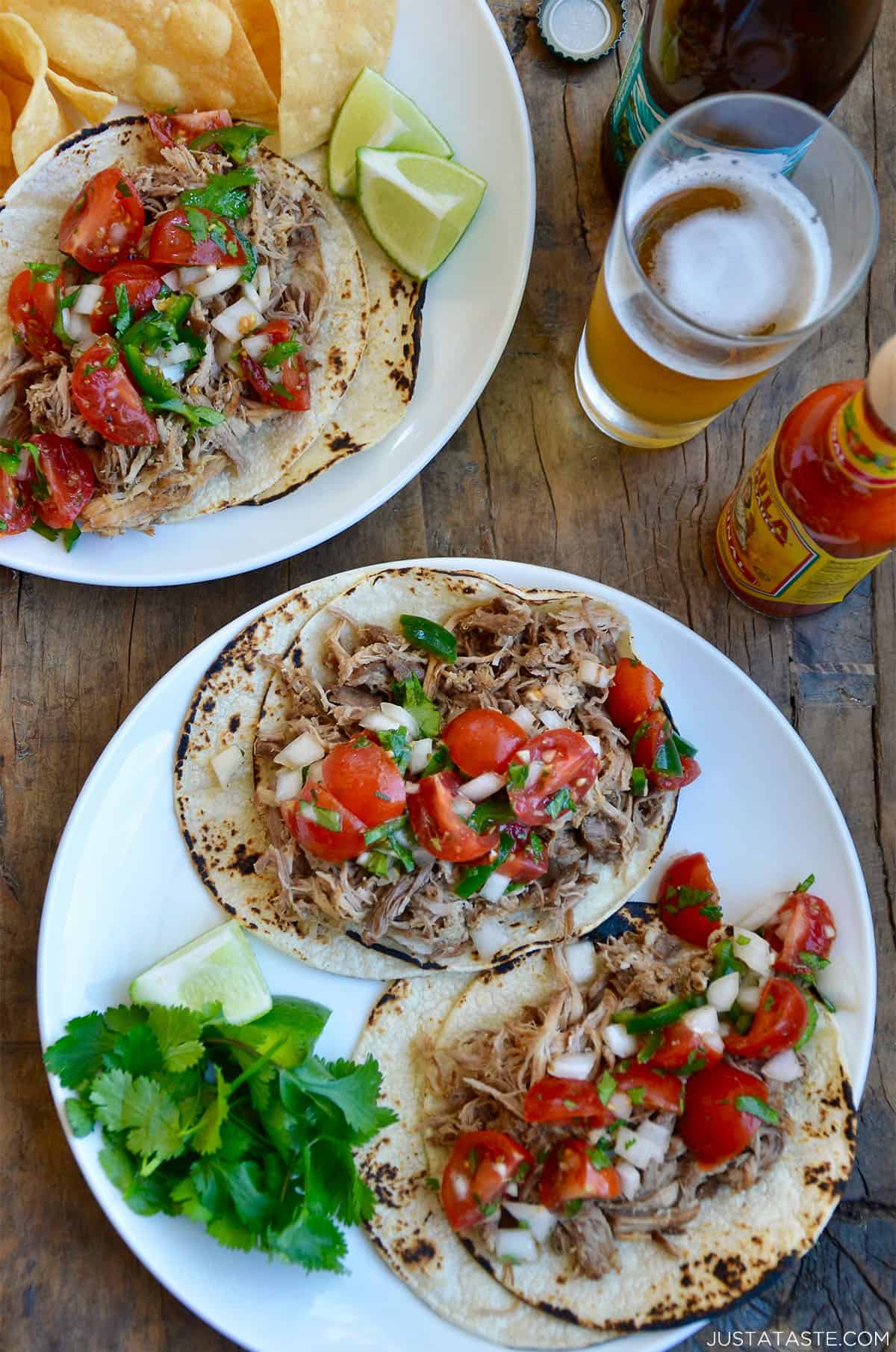 Traditional carnitas (Mexican pulled pork) atop corn tortillas with chopped cilantro and pico de gallo. A glass of beer and hot sauce are nearby.