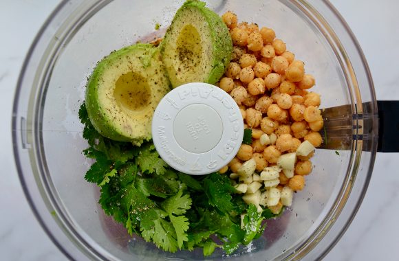 A clear food processor bowl containing avocado, chickpeas, garlic and herbs