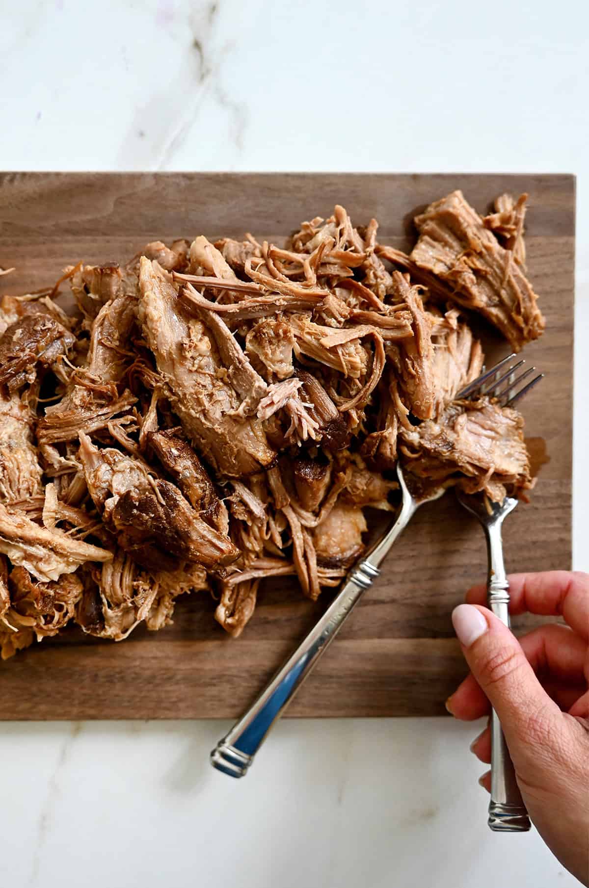 Using two forks to gently shred pork on a cutting board.