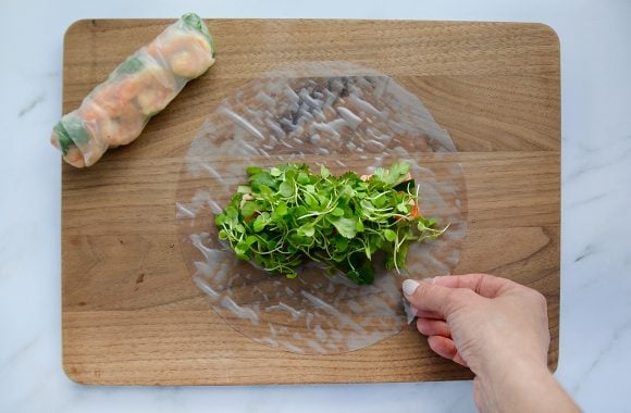 A hand placing micro greens atop a rice paper sheet with a completed Thai spring roll next to it 