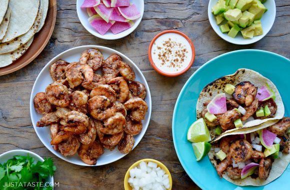Plates and bowls containing spicy shrimp tacos, spicy shrimp, tortillas, sour cream, avocado and cilantro