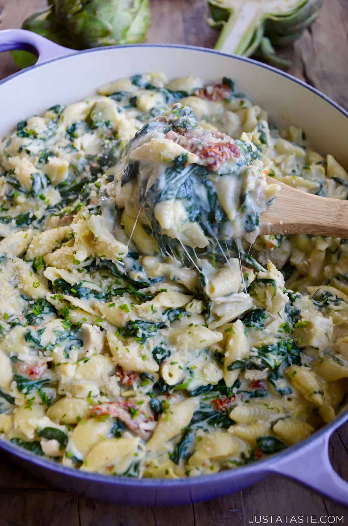 A large wooden spoon digs into a stockpot containing creamy spinach pasta with artichokes, sun-dried tomatoes and chicken.
