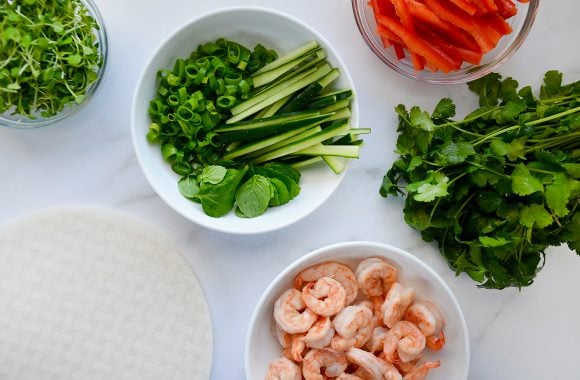 Rice paper sheets and small bowls containing micro greens, english cucumber with scallions, red bell pepper, cilantro and shrimp