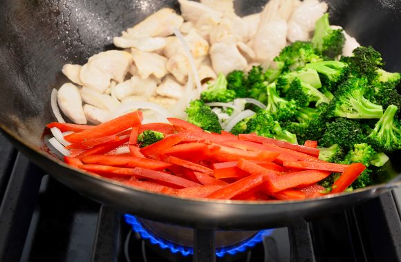 Wok containing sliced red bell pepper, broccoli florets and chicken over oven burner