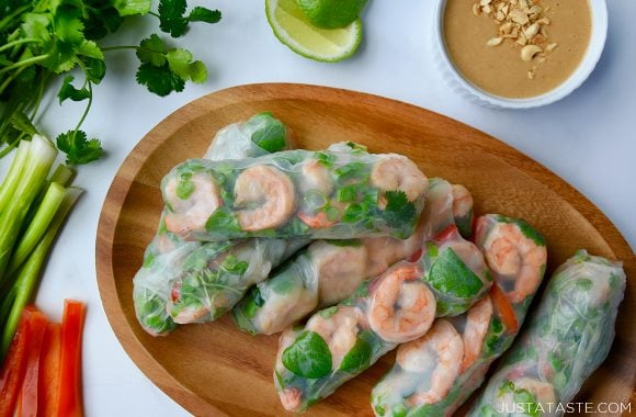 A wood serving plate containing Thai Spring Rolls with peanut sauce in a small bowl, limes and veggies around it